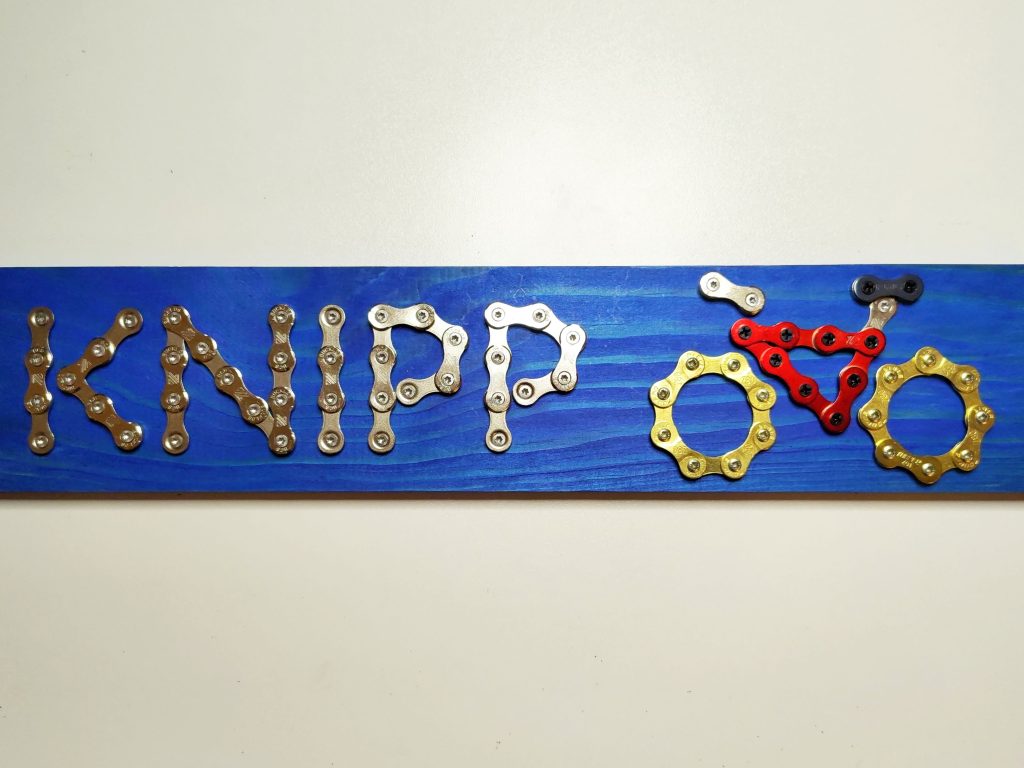 Türschild, welches einen Nachnamen und daneben ein Fahrrad aus Fahrradkettenteilen aufgeschraubt auf einem blauen Holzbrettchen zeigt.