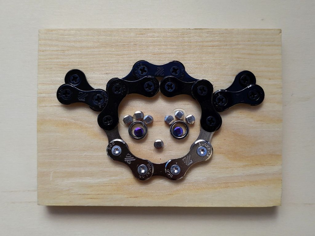 Motivschild welches ein Mädchen mit schwarzen Haaren zeigt. Das Motiv ist aus Fahrradkettenteilen zusammengesetzt, aufgeschraubt auf einem Holzbrettchen.